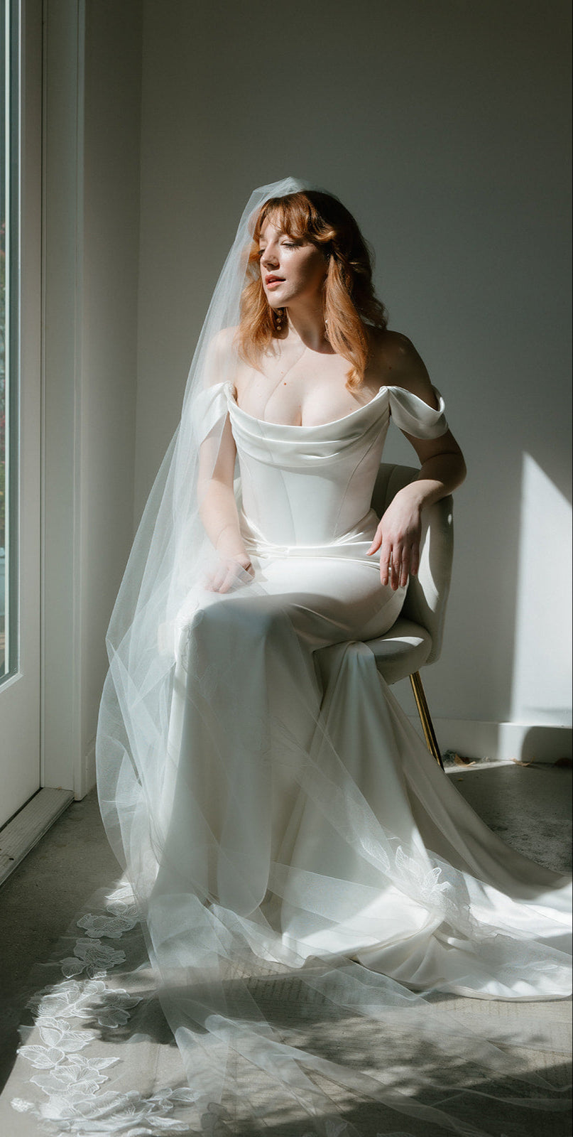 Woman in a Corston Couture Temptress wedding dress sitting by a window with sunlight streaming in onto Aunyalune Reverie veil.