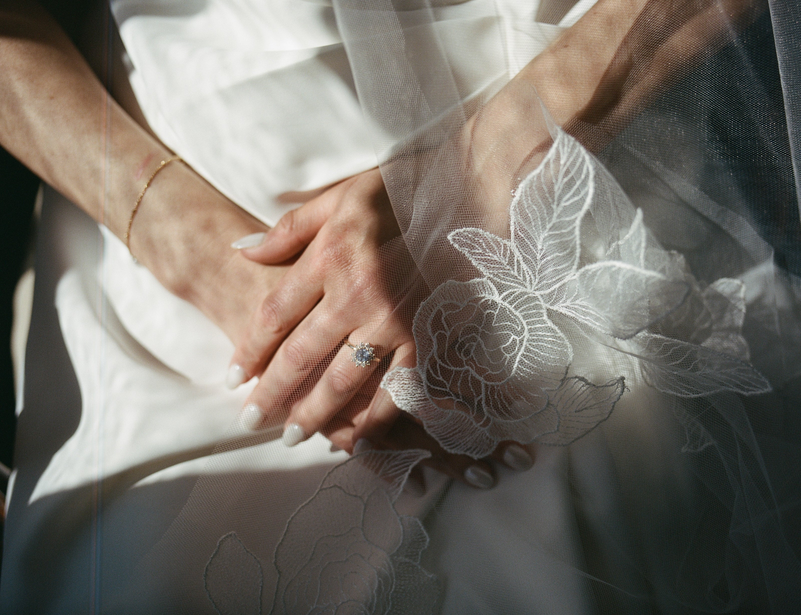 Close-up of hands holding each other with a wedding ring, surrounded by french embroidered peonies botanical lace veil.